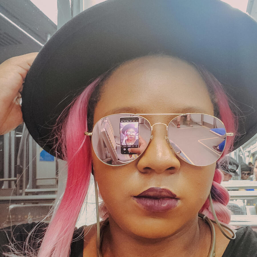 Self portrait of Imani J Dean a Black femme presenting adult. They are wearing reflective sunglasses that you can see the phone they're using to take the photo in. They have a black hat on. Their hair is styled in two pink braids. They have a dark purple/m